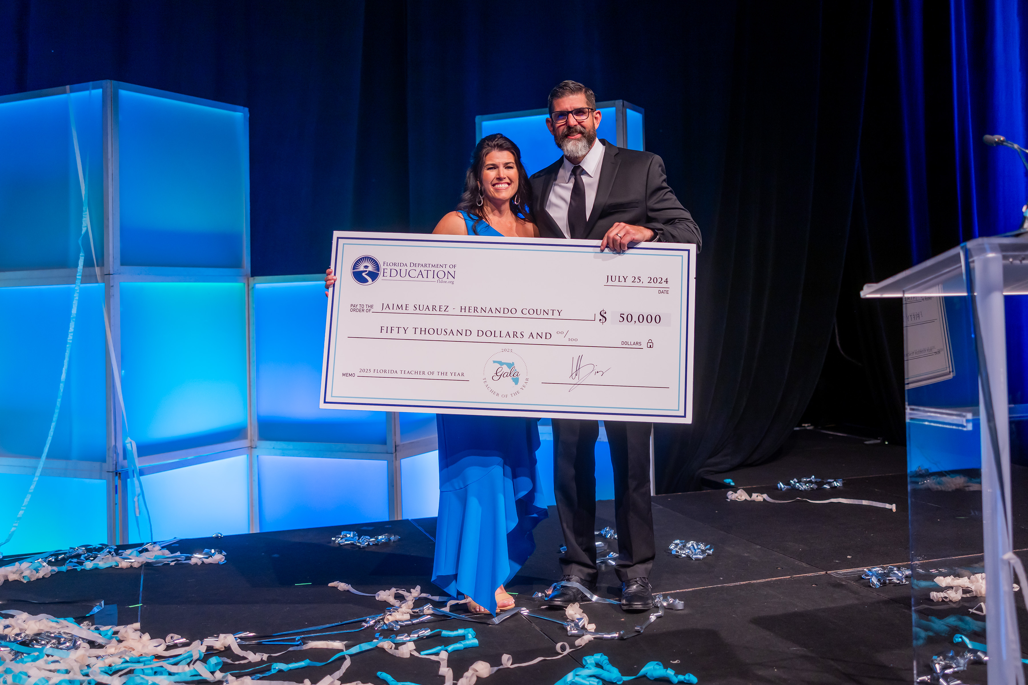 2025 Teacher of the Year Jamie Suarez holding up check with Commissioner of Education Manny Diaz, Jr.